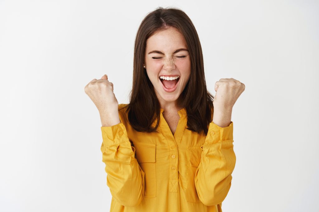 Young satisfied woman winning prize and celebrating, making fist pump and screaming of joy, triumphing on white background